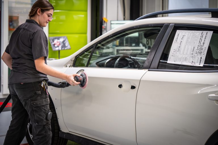 Woman using a polishing tool on a white vehicle in an auto body shop, demonstrating professional autobody repair techniques for collision restoration.