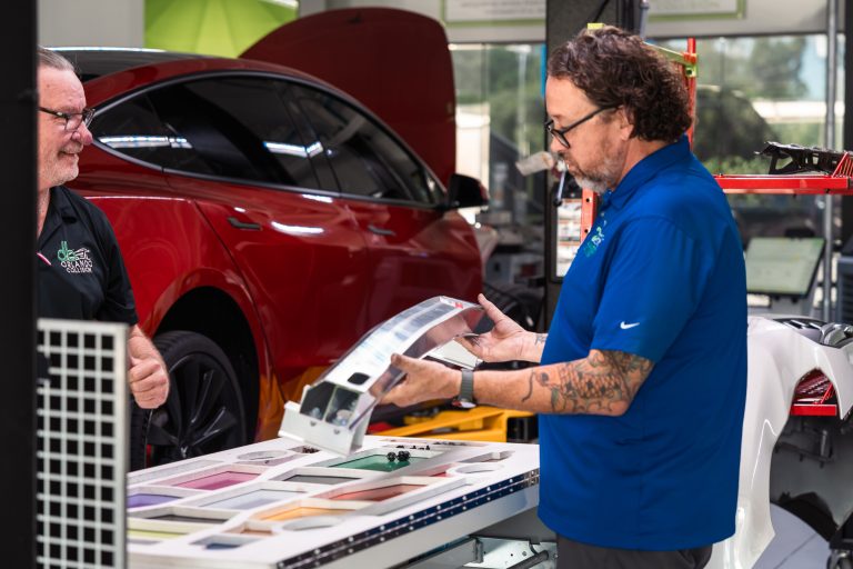 Two auto body repair technicians discussing a vehicle panel color match in an auto body shop, with a red car visible in the background and various paint samples displayed on a table.