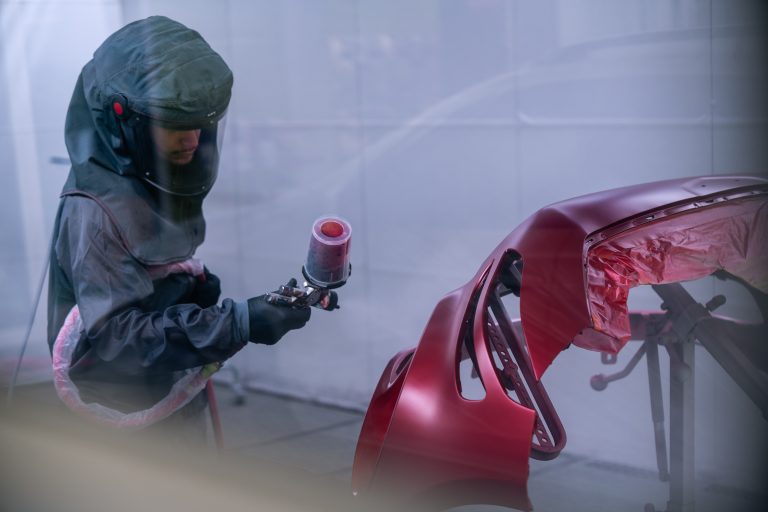 Auto body technician using a spray gun to apply red paint on a vehicle part in a professional collision repair shop, showcasing high-quality autobody repairs and refinishing services.